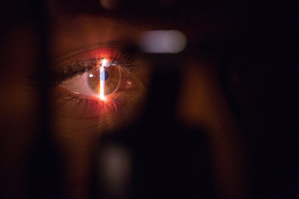 Close-up of a human eye undergoing an eye exam with a bright light shining directly into the pupil, highlighting the details of the iris and reflection on the cornea.