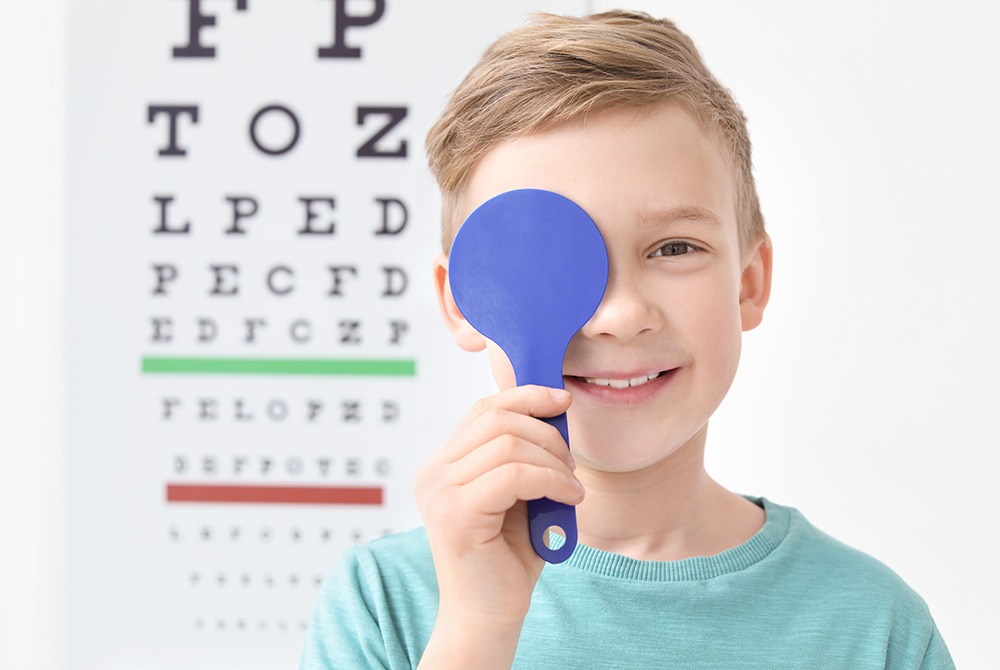 A smiling boy covers one eye with a blue occluder during an eye exam, standing in front of a blurred vision chart.