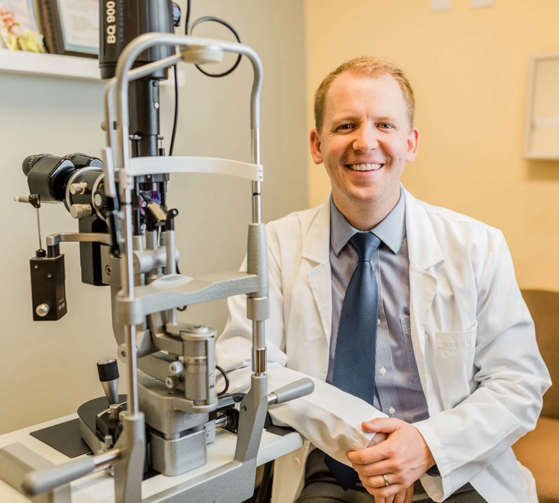 A smiling male optometrist in a white coat sits beside a slit lamp eye examination machine in a well-lit clinic room.