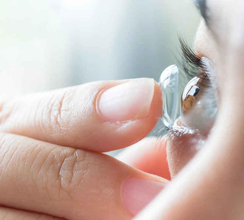 A close-up of a person’s hand inserting a clear contact lens onto their eye, with eyelashes and part of their face visible in the background.